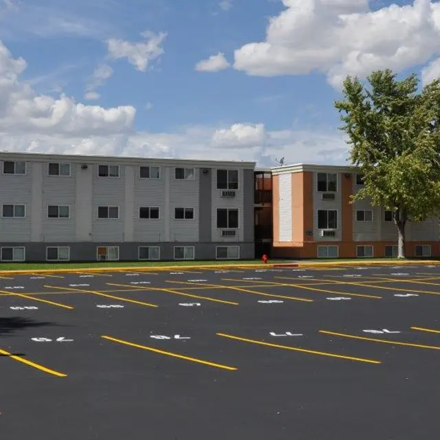 A view of an apartment complex next to a spacious parking lot, featuring several empty parking spaces and trees in the background under a partly cloudy sky.