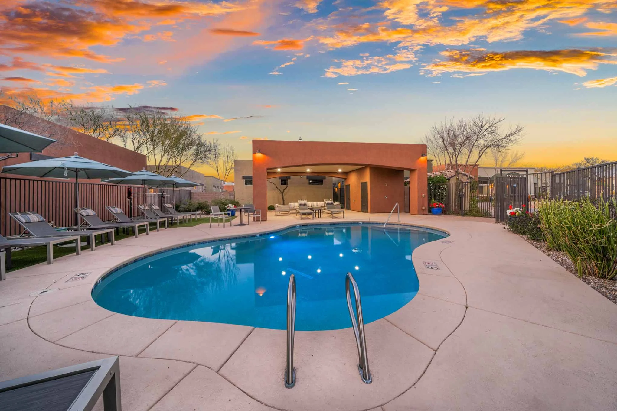 Avilla Marana A serene poolside scene at sunset, featuring a clear blue pool, lounge chairs, and atmospheric clouds in the sky.