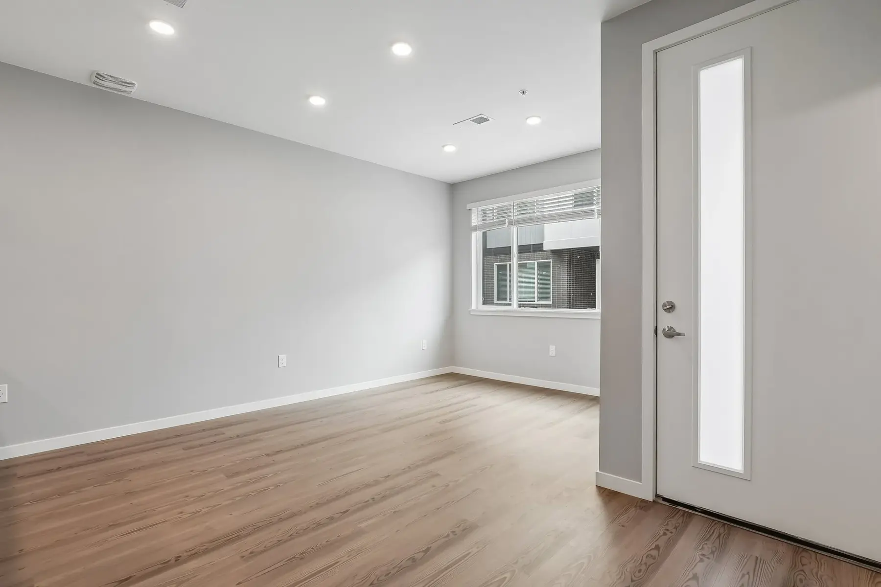 Empty Apartment Room A clean, empty room with light gray walls and wooden flooring, featuring a large window and a modern door.