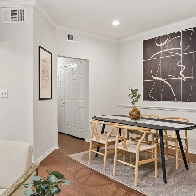 A modern dining area featuring a round black dining table surrounded by light wood chairs. A large abstract art piece adorns the wall behind the table, while a small decorative plant sits on the table. The space has a warm, inviting ambiance with neutral colors.