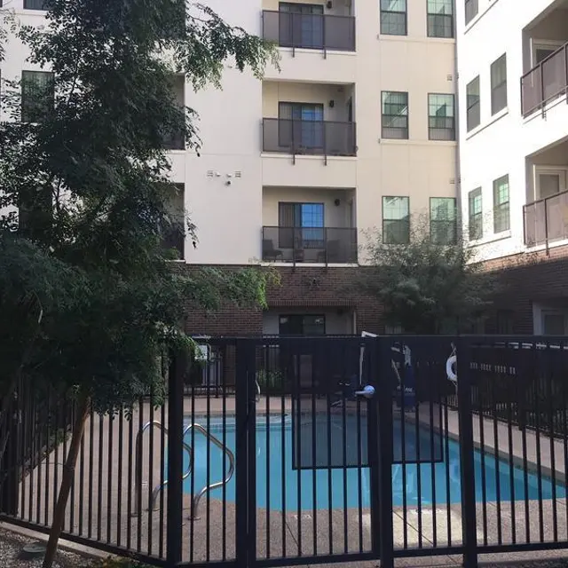 A view of a rectangular swimming pool enclosed by a black metal fence, surrounded by landscaped greenery, located in an apartment complex with multiple floors.