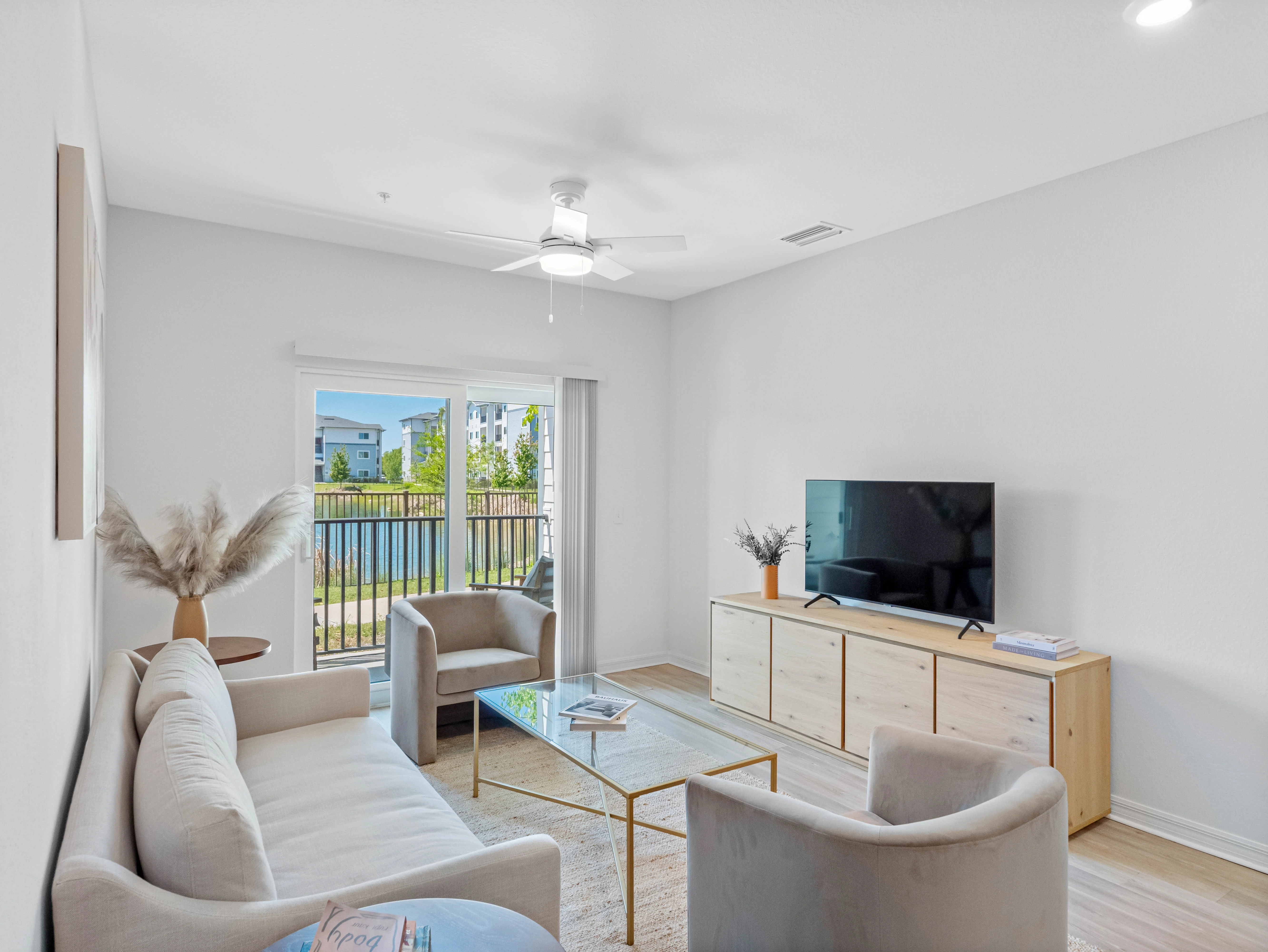 A bright and modern living room featuring a beige sofa, two light gray armchairs, a glass coffee table, and a TV on a wooden console. The room has a ceiling fan and large windows with a view of the outdoors.