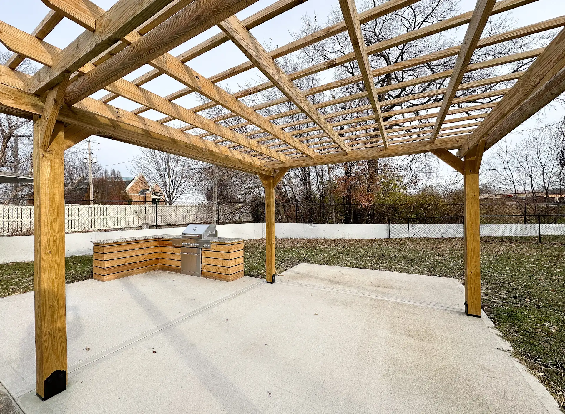View of a wooden pergola structure with an open roof over an outdoor grilling area, featuring a stone grill counter.