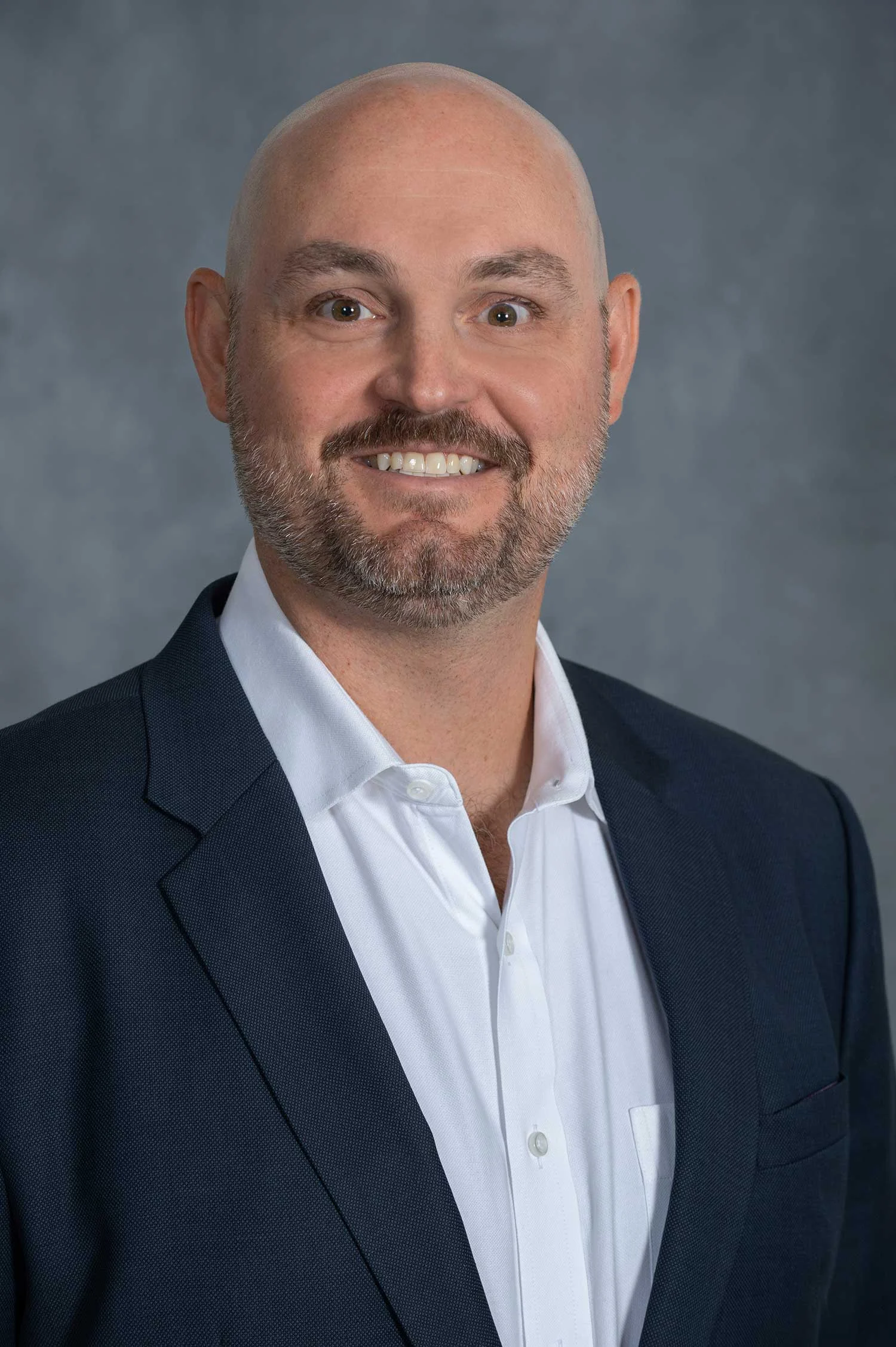 A smiling man with a bald head and a beard, dressed in a dark suit jacket over a white shirt, against a gray background.