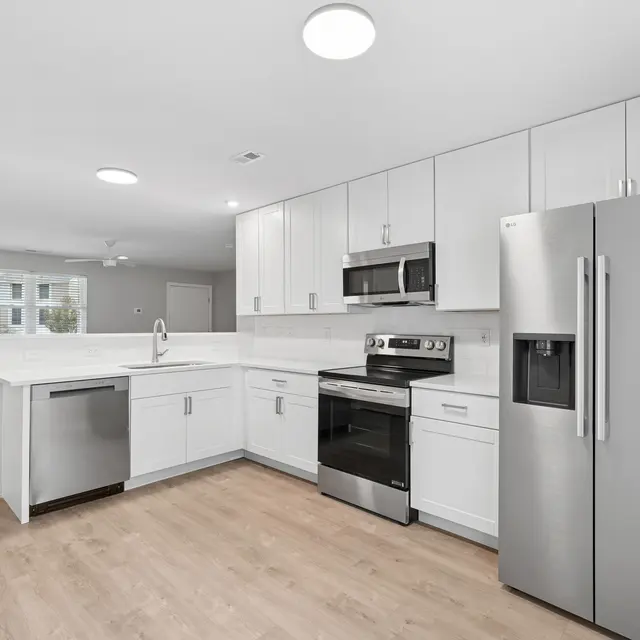 A modern kitchen featuring stainless steel appliances, including a refrigerator, oven, and dishwasher. The cabinetry is white with a clean finish, and the countertops are light-colored. The space has a neutral color scheme with wooden flooring and ample natural light coming through a window.