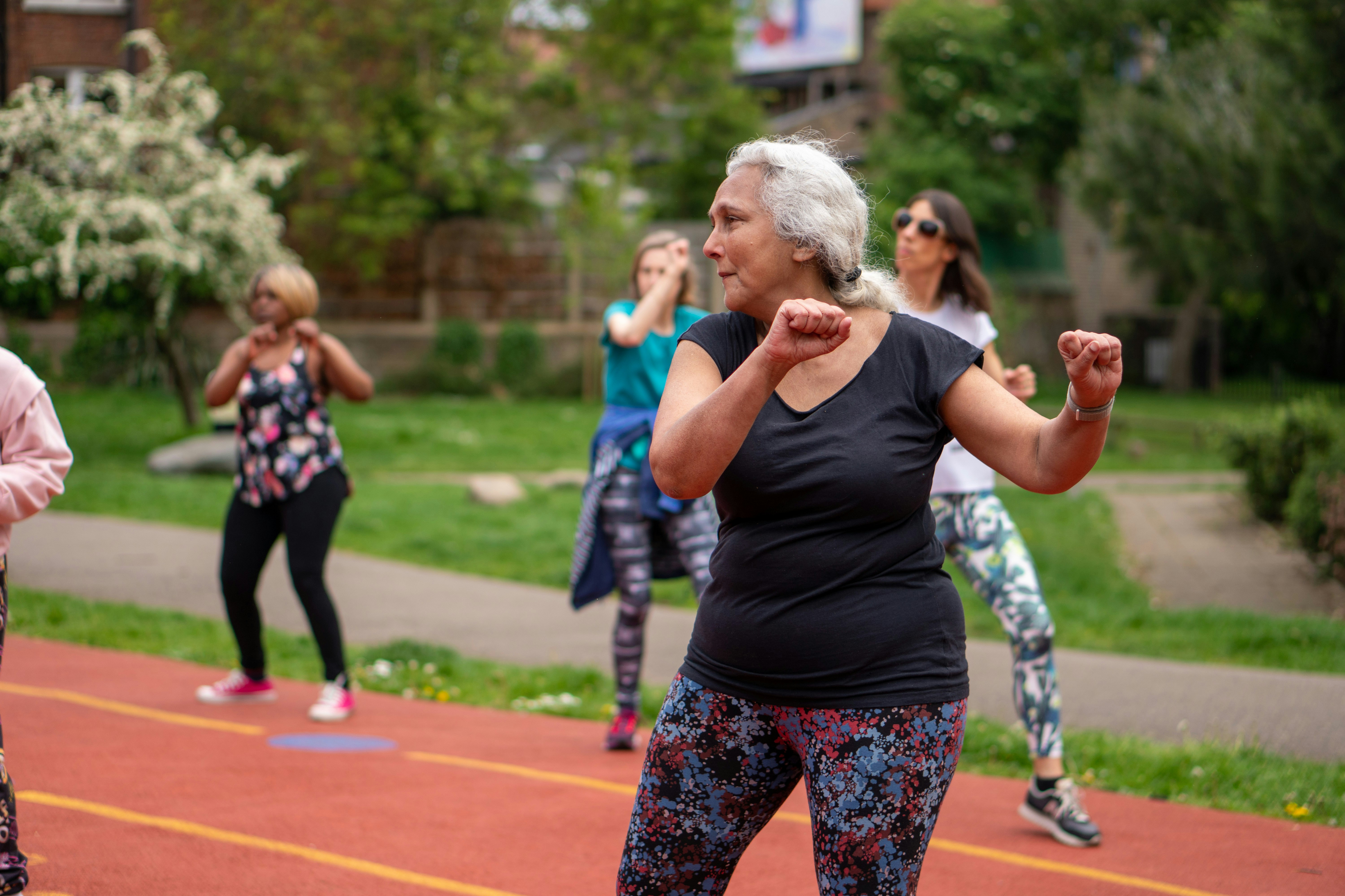 Outdoor Zumba Class in the Park A group of diverse individuals participating in a Zumba class outdoors, with a focus on an older woman in a black top and colorful leggings leading the movements. The setting is a vibrant park with greenery in the background.