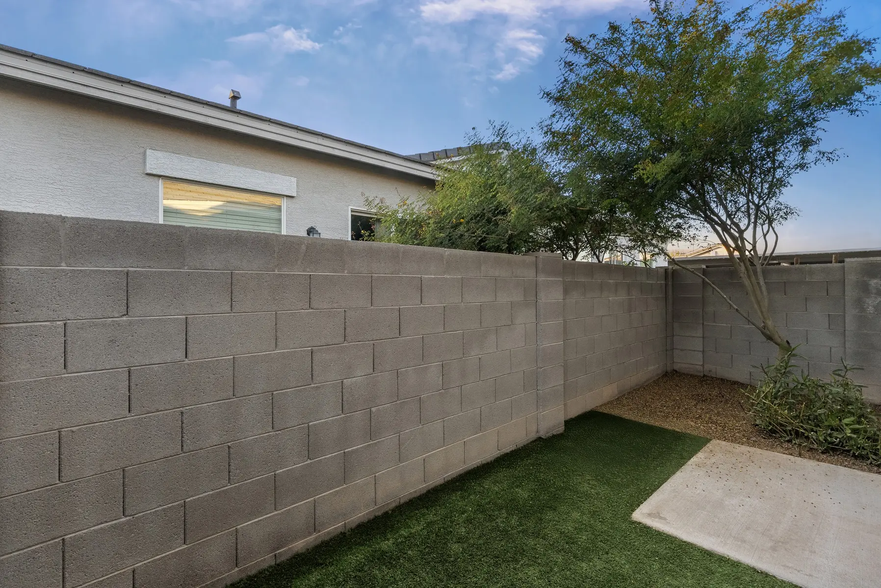 A backyard view featuring a gray block wall, a small patch of grass, and a few shrubs under a blue sky.