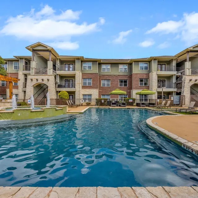 A view of a luxury apartment complex with a swimming pool and lounge area. The pool is surrounded by stone and has lounge chairs nearby. In the background, there are multiple levels of the apartment building with balconies and greenery.