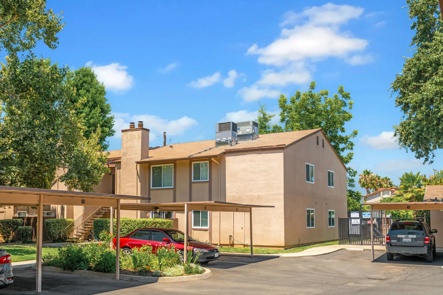 A residential apartment complex with two floors, featuring trees and blue sky in the background. Cars are parked under a shade structure in the foreground.