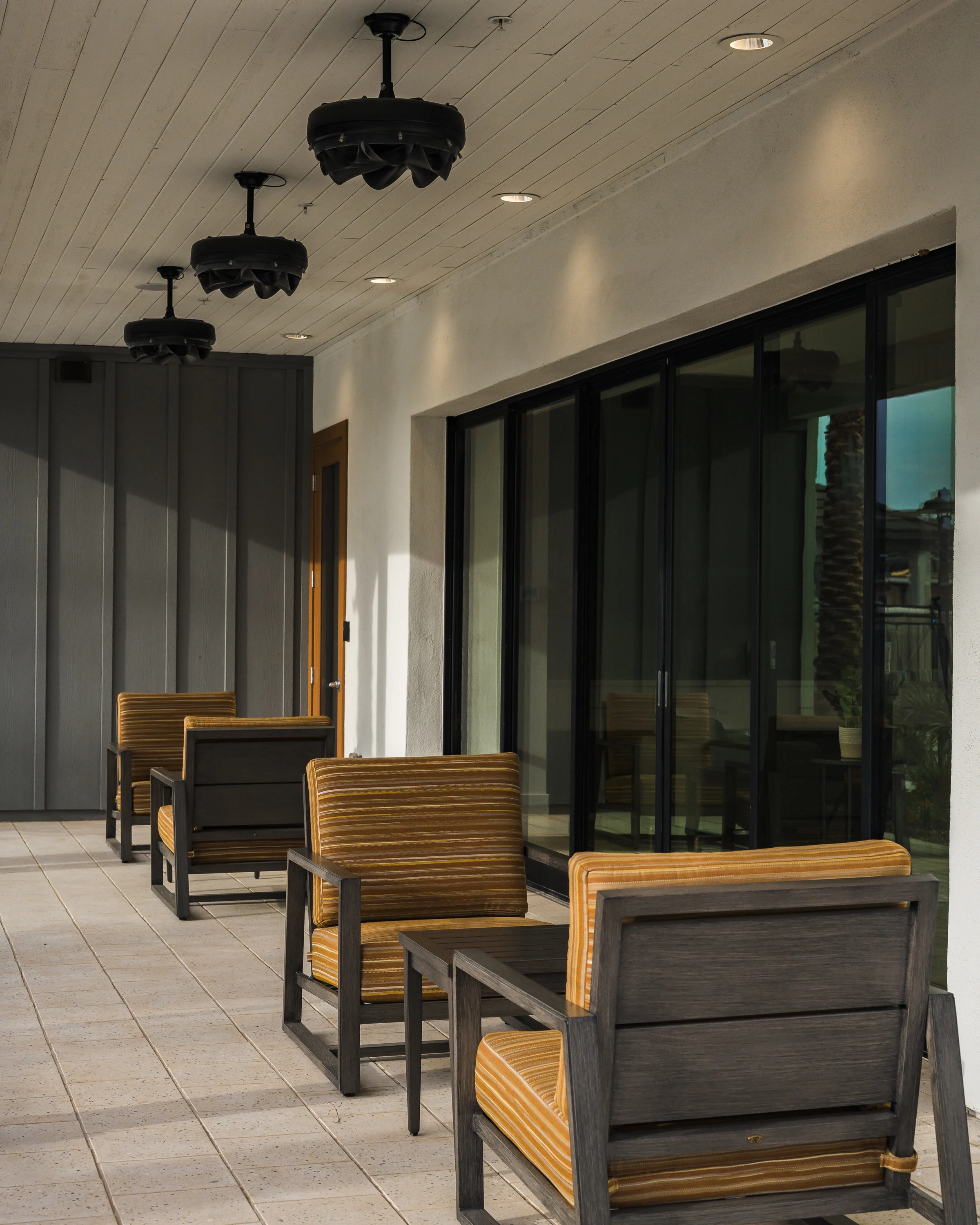 Peralta Vista A modern porch area featuring wooden chairs with orange striped cushions and large glass windows. The ceiling has unique black pendant lights, and the floor is tiled with light-colored tiles.