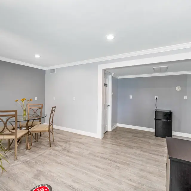 A spacious, modern living area featuring a small dining table with four chairs, a potted plant, and a black refrigerator. The room has light-colored walls and wooden flooring, creating a bright atmosphere.