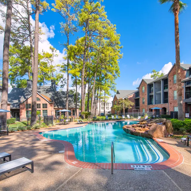 A well-maintained apartment complex pool area with lounge chairs, surrounded by trees and buildings.