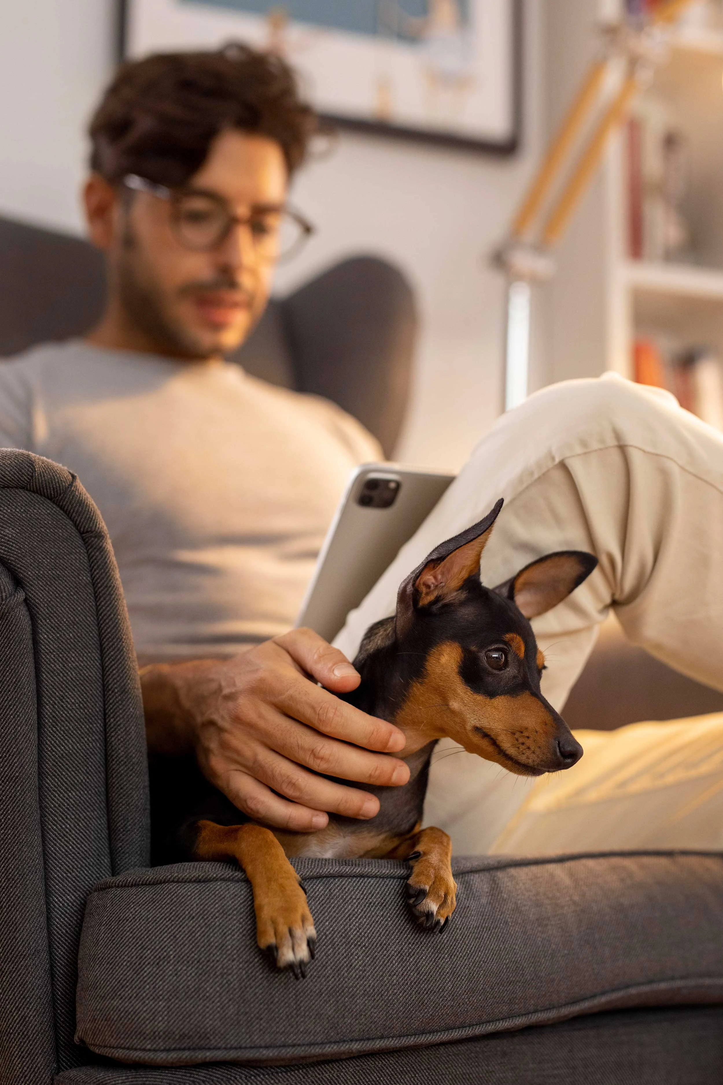 A man sitting on a couch, reading an iPad, with a small dog resting beside him.