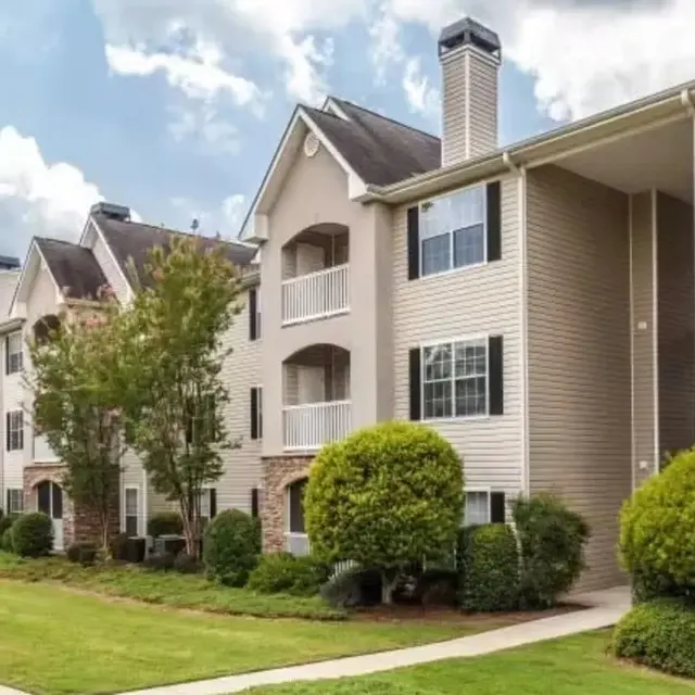 Exterior view of a multi-unit residential building with a manicured lawn, trees, and walkways under a partly cloudy sky.