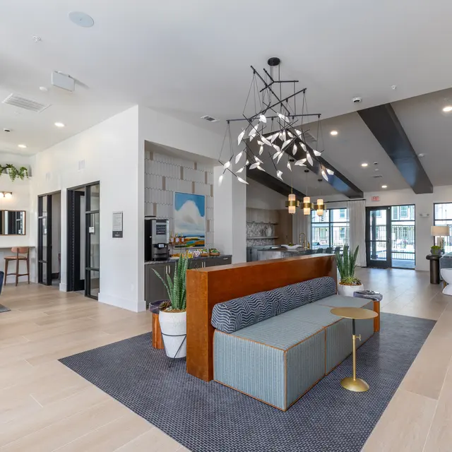 A modern lobby interior featuring a cozy seating area with a wooden and upholstered couch, a sleek light fixture, and open spaces leading to glass doors. Decor includes potted plants and stylish furnishings.