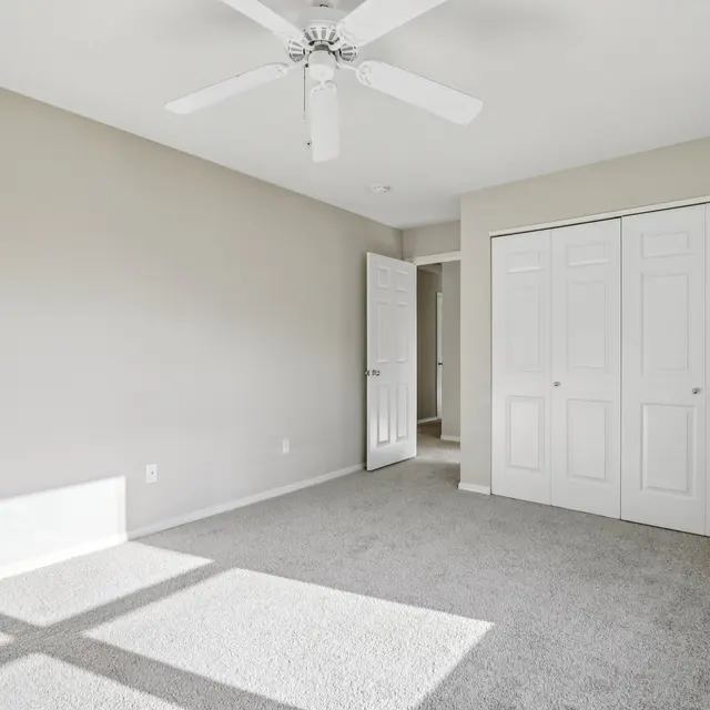 A spacious empty bedroom with light-colored walls and a ceiling fan, featuring two large closet doors and a doorway leading to another room.