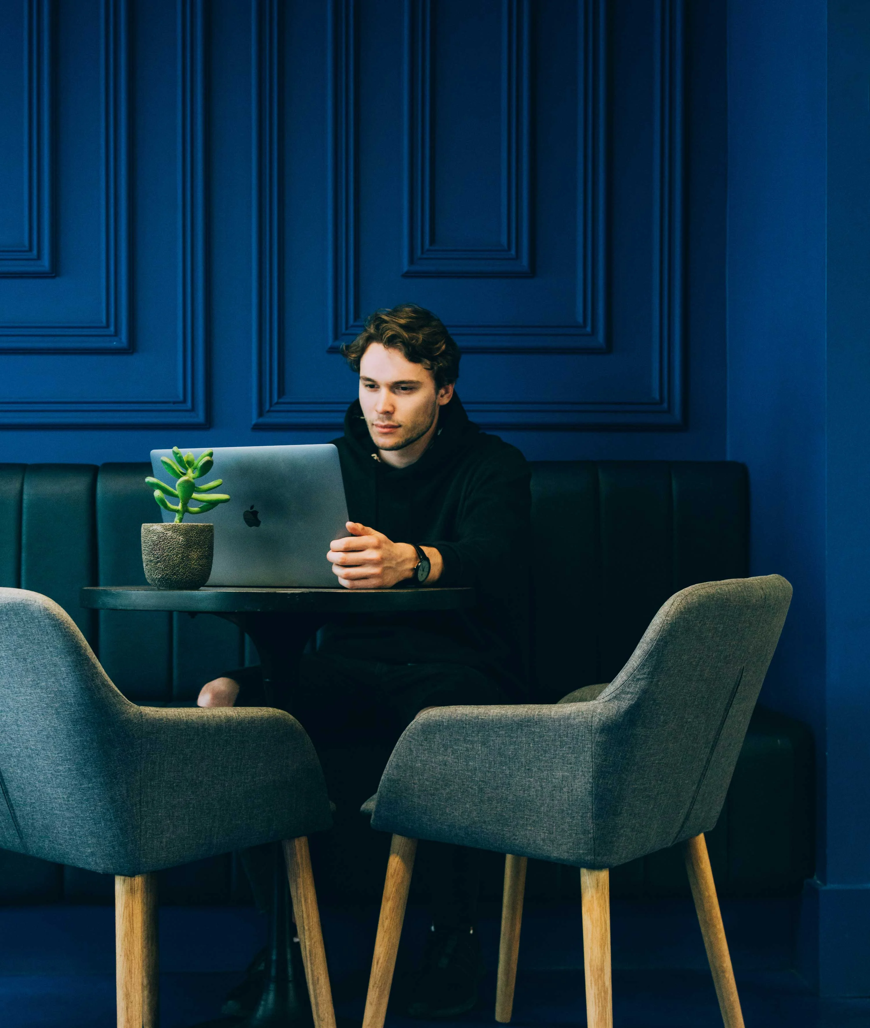 A young man sitting at a table with a laptop, surrounded by gray chairs and a small plant, against a dark blue wall.