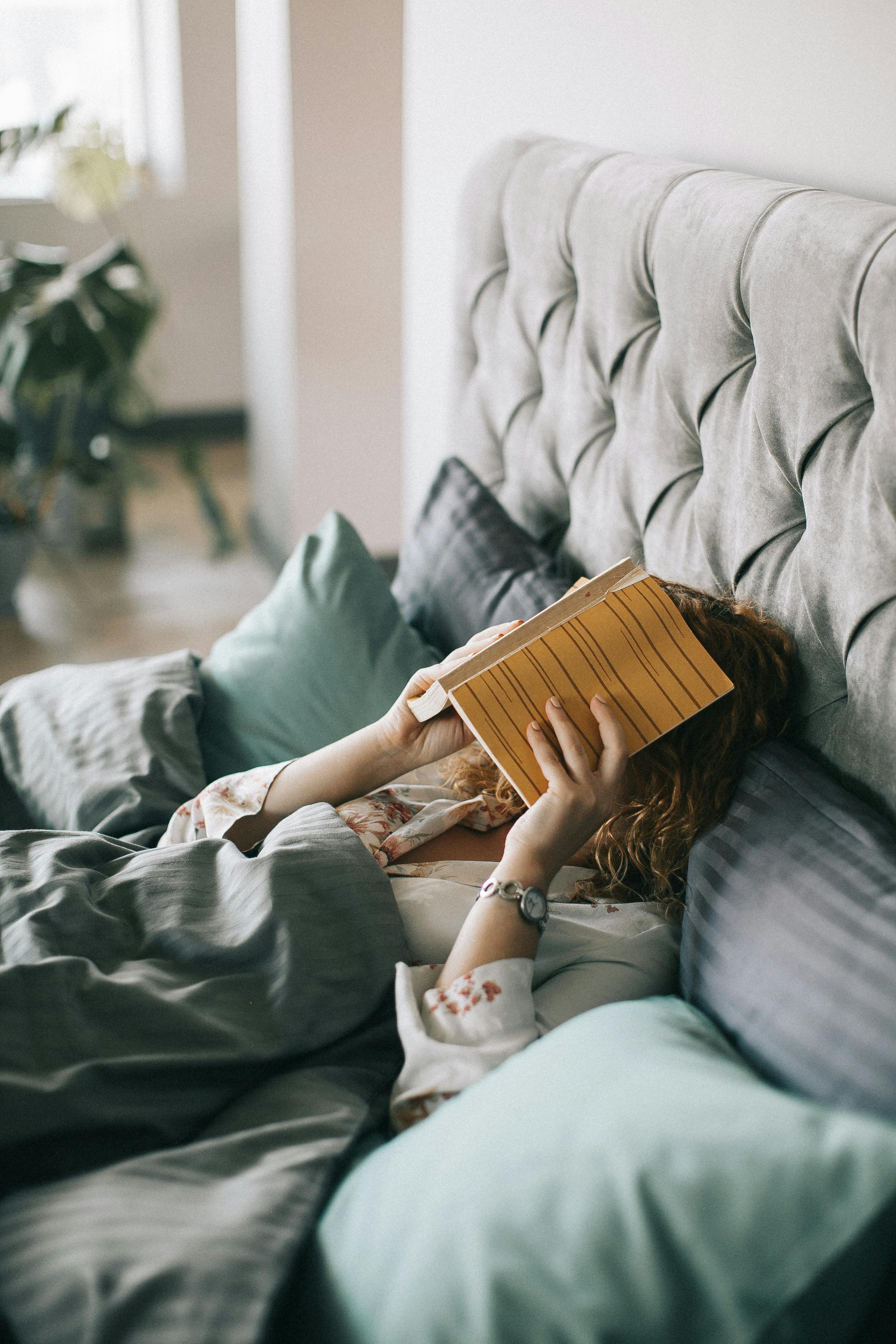 A woman lying in bed covered with blankets, holding a book in front of her face, surrounded by pillows in a cozy setting.