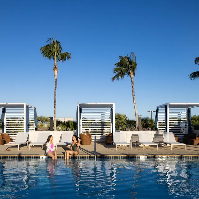 Two women relaxing at a poolside seating area under palm trees, with luxurious cabanas in the background and a clear blue sky.