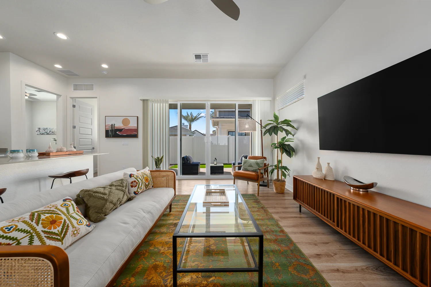 A stylish modern living room featuring a white sofa with colorful cushions, a glass coffee table, a wooden TV stand, and a large wall-mounted TV. The room has large sliding glass doors that open to a backyard, with indoor plants and a decorative rug on the floor.