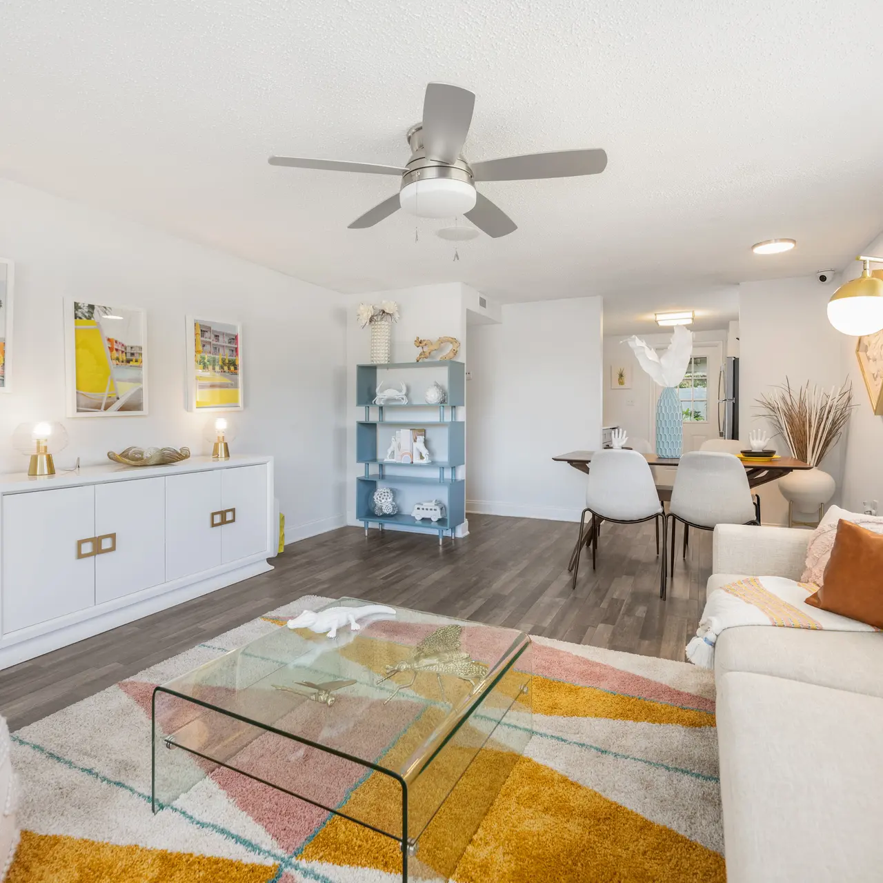 A modern living room featuring a white sofa with orange cushions, a colorful area rug, a glass coffee table, and a stylish shelving unit with decorative items. There are dining chairs and a table visible in the background as well as wall art.