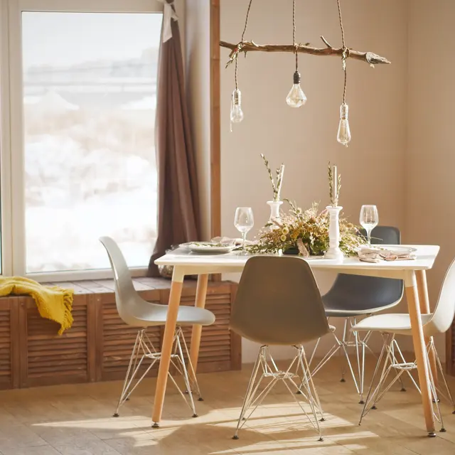 A cozy, modern dining area featuring a table with decorative plants, glassware, and hanging light bulbs. The room has large windows with a view of a snowy landscape.