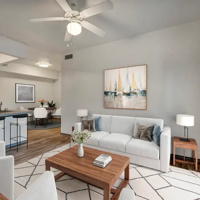 A modern living room featuring a white sofa with decorative pillows, a wooden coffee table, and a ceiling fan. The walls are light-colored and adorned with a nautical-themed artwork. In the background, there's a dining area with white chairs and a glimpse of a kitchen.