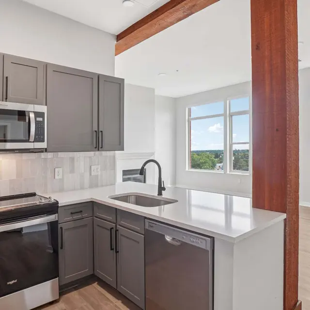 Modern Kitchen Interior A modern kitchen featuring dark cabinetry, stainless steel appliances, and a white countertop. A dishwasher is visible, and there is a window with a view outside. The adjacent space has large windows, allowing natural light in, and includes a wooden support beam.
