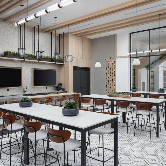 A modern kitchen area featuring long tables with high stools, wood and tile finishes, and large windows letting in natural light.