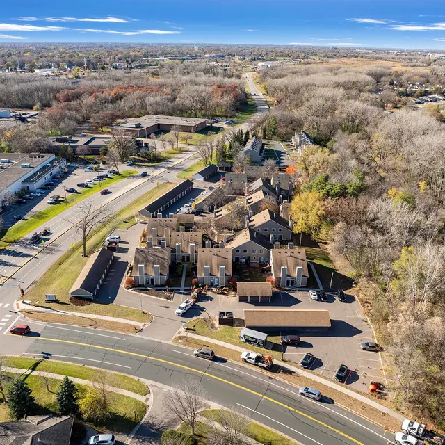 Aerial view of a residential area featuring multiple buildings and a curved road. Surrounding landscape shows trees with autumn colors and a clear blue sky.