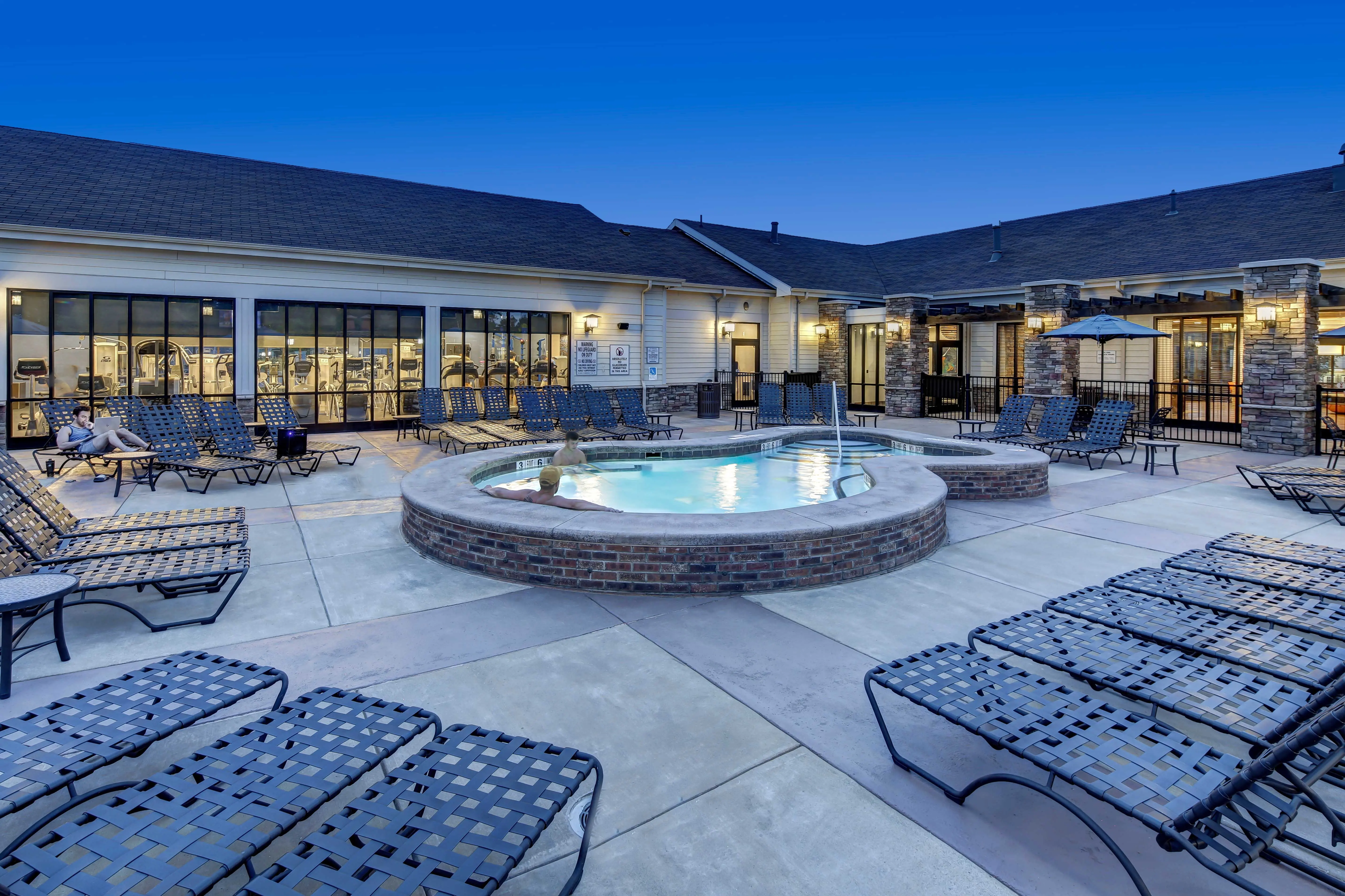 An outdoor swimming pool area featuring a hot tub surrounded by lounge chairs under a twilight sky.