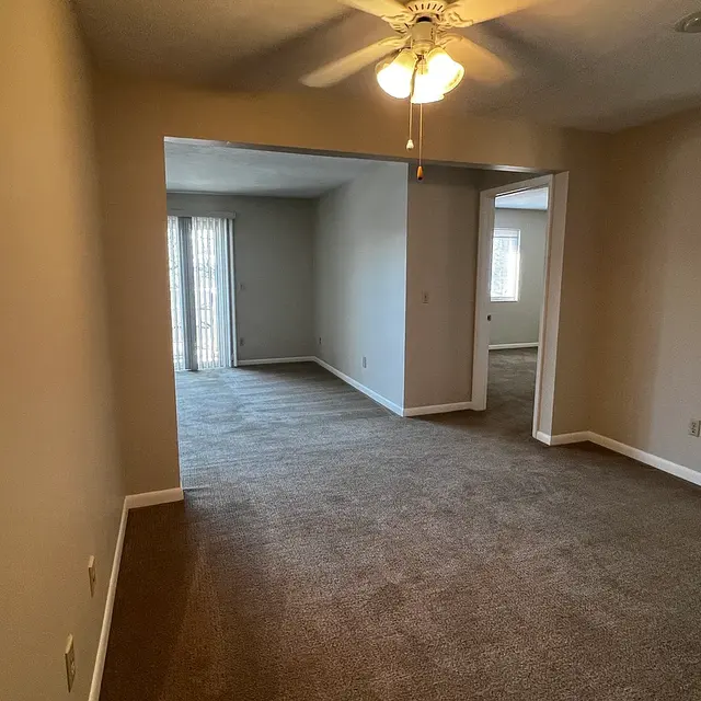 A spacious living room area with beige walls and brown carpet, featuring a ceiling fan and an opening leading to another room with sliding glass doors in the background.