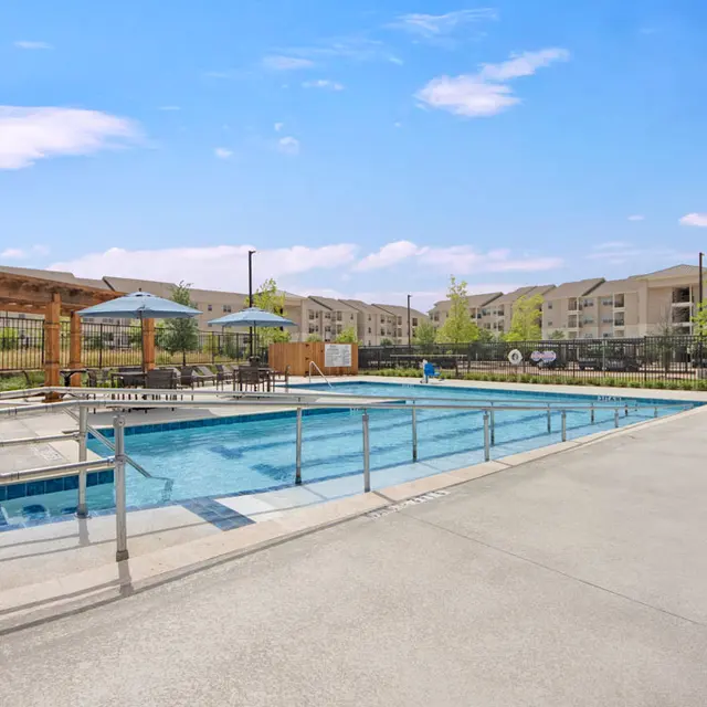Apartment Complex Pool Area A bright and sunny pool area at an apartment complex. The pool features a railing, surrounded by concrete and a fence. There are shaded seating areas nearby and buildings in the background under a clear blue sky.