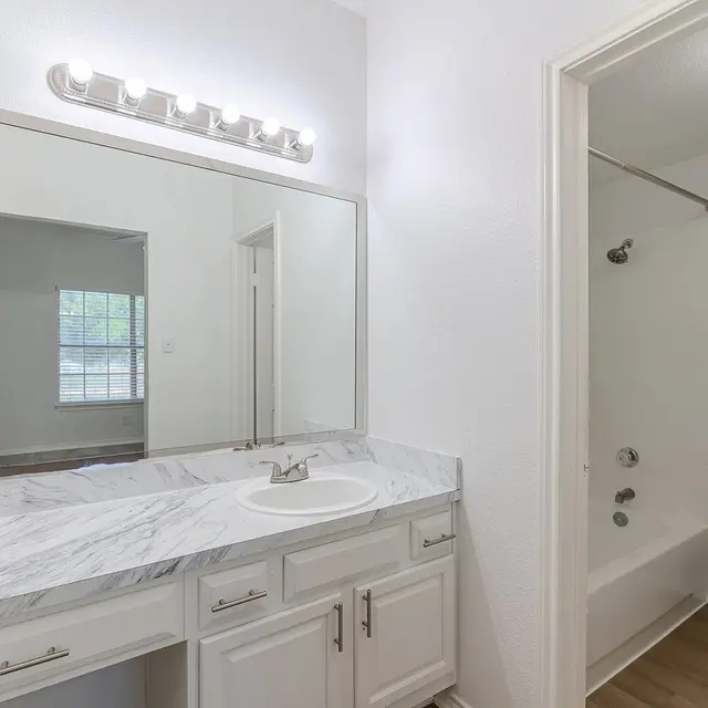 A modern bathroom featuring a dual sink vanity with a marble countertop and a large mirror. The space has neutral-colored walls and is well-lit by a vanity light above the mirror. On the side, there's a doorway leading to a small bathroom area with a bathtub, shower, and toilet, all maintained in a clean and contemporary style.