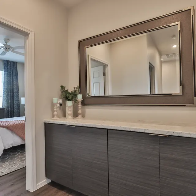 A modern hallway with a decorative mirror and a console table. In the background, a well-lit bedroom is visible with a bed and large window.