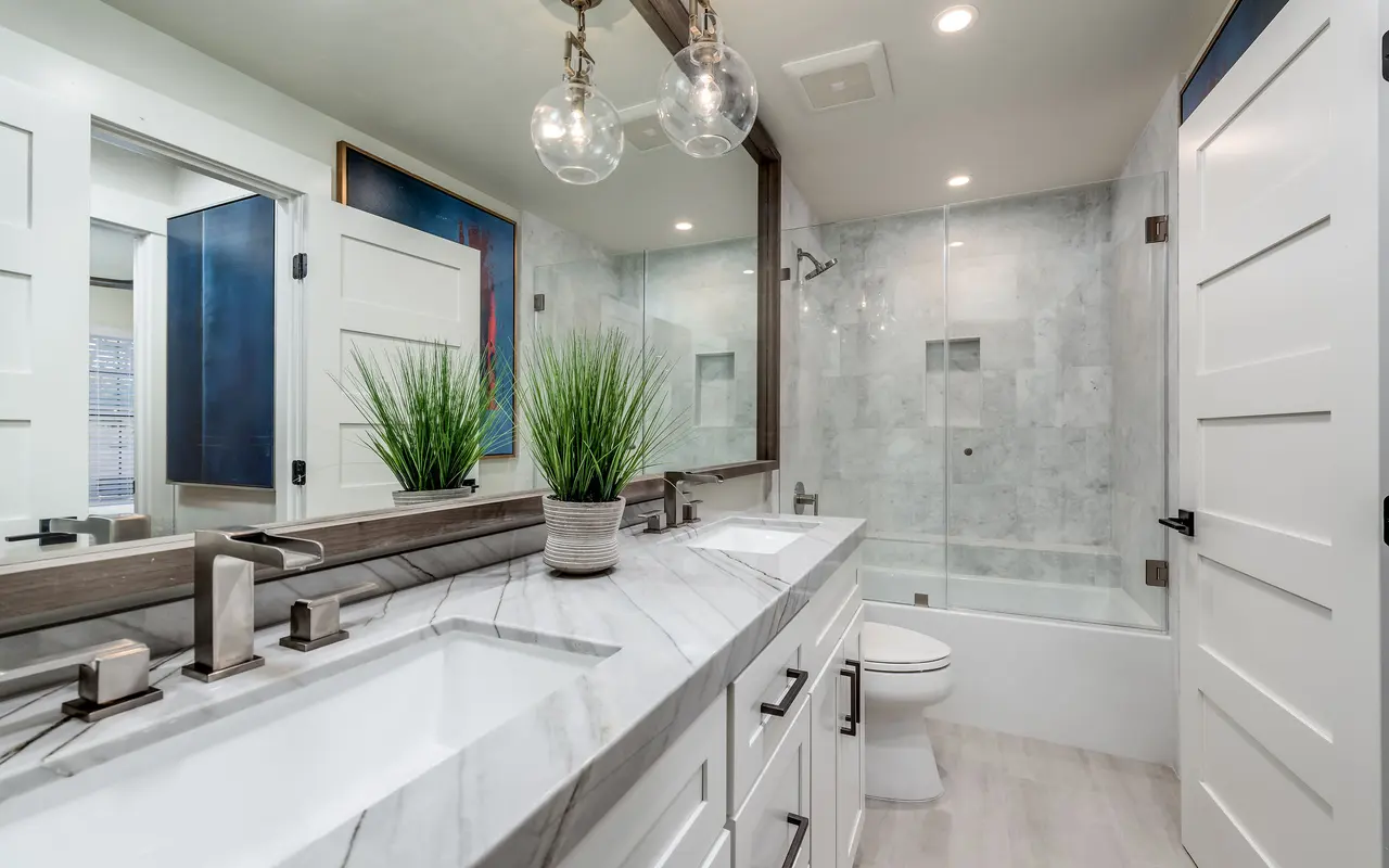 A modern bathroom featuring a double vanity with a marble countertop, two sinks, and faucets, complemented by a potted plant. There is a glass shower enclosure in the background, alongside a toilet, with large mirrors and bright lighting.