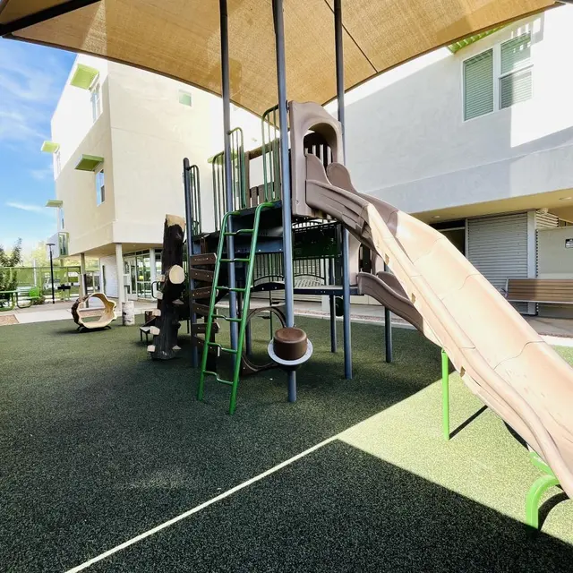 A children's playground featuring a brown slide, climbing structure, and soft green flooring. The background consists of modern buildings with large windows and covered areas for shade.