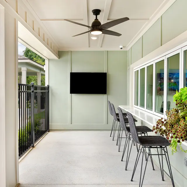 A modern patio space featuring a light green wall, a ceiling fan, black chairs, a television mounted on the wall, and a potted plant arrangement.