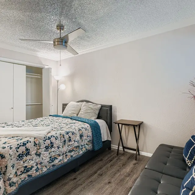 A cozy bedroom featuring a bed with a floral-patterned comforter and blue accents, a couch, and a ceiling fan, with light wood flooring and a modern aesthetic.