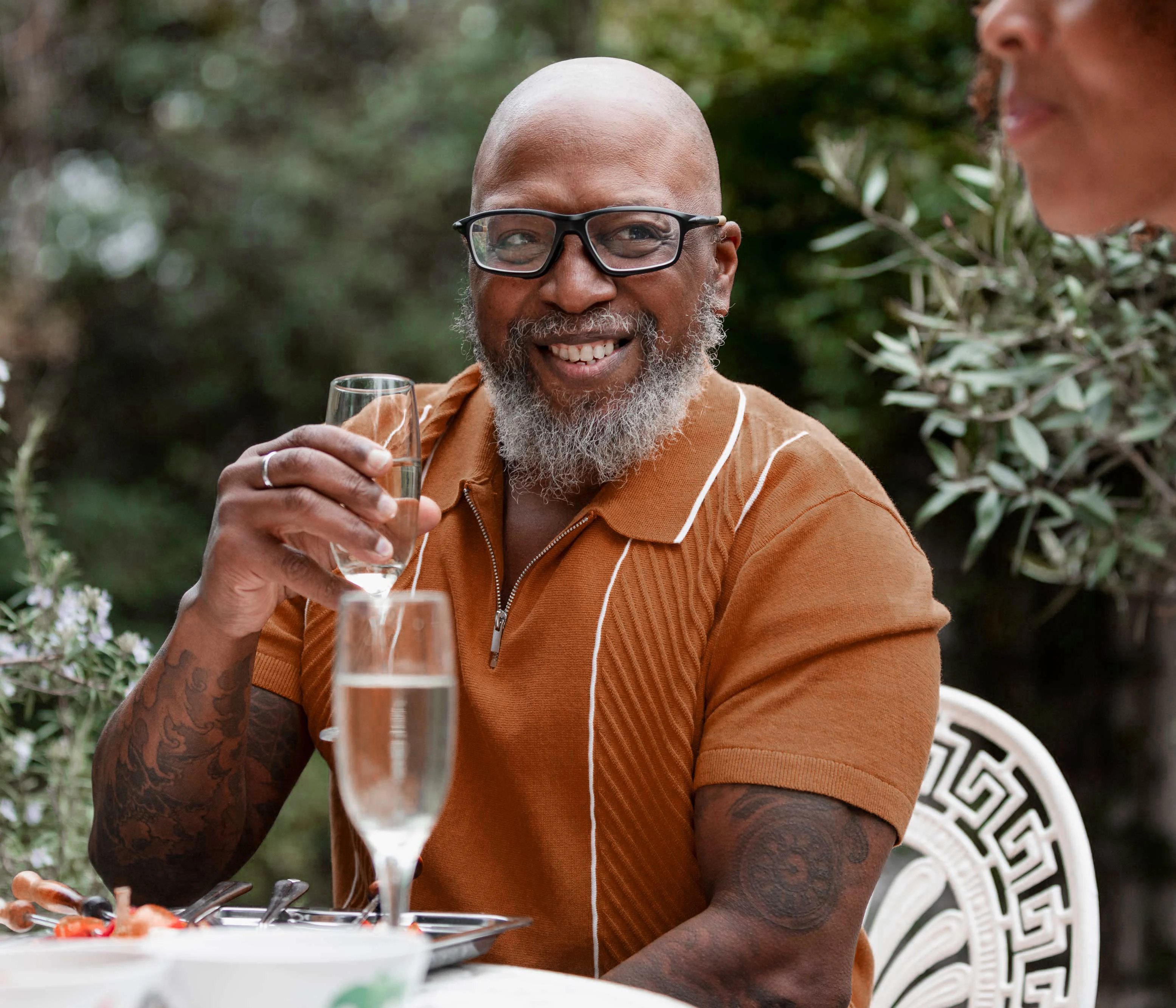 A man with a beard and glasses smiles while holding a glass of champagne at a dining table, with greenery in the background.