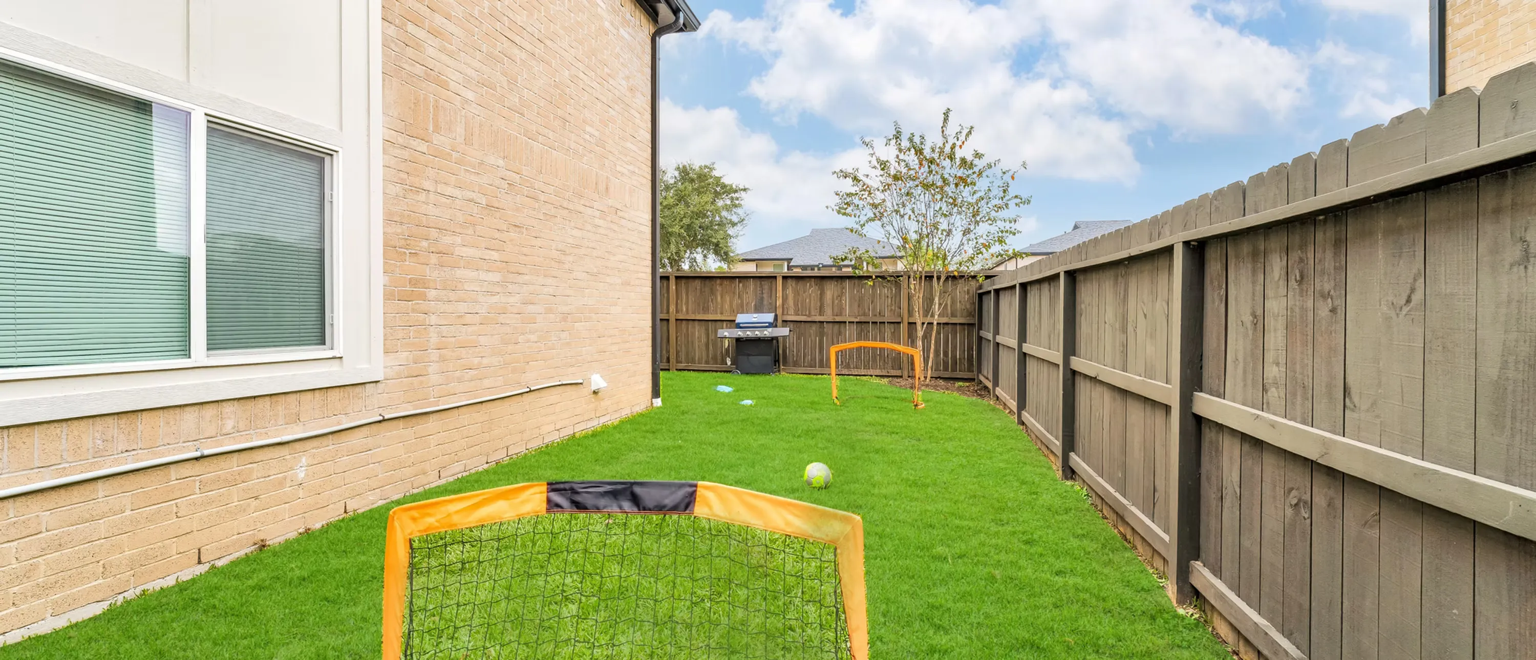 View of a backyard with artificial grass, a grill, and two small goalposts