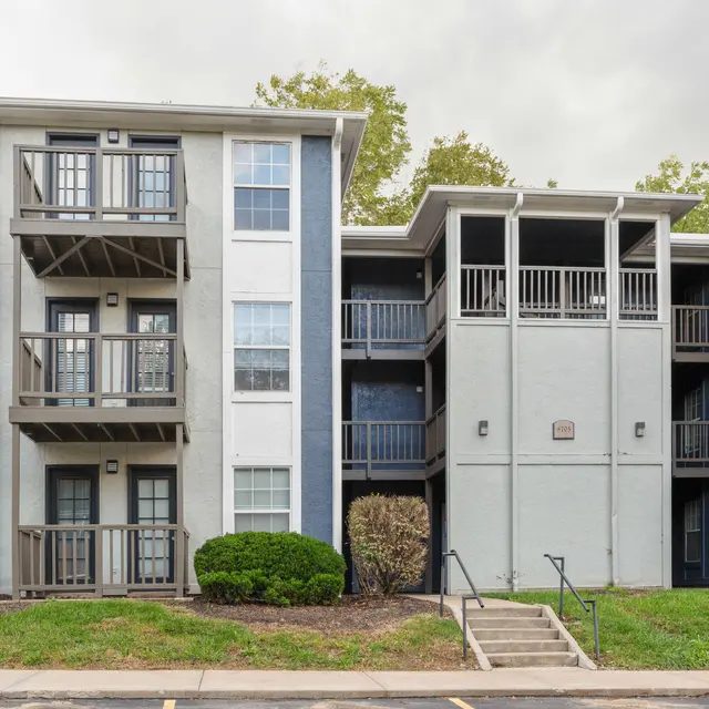 A multi-story apartment complex featuring three sections with balconies, greenery in front, and a cloudy sky.