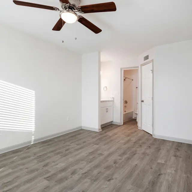 A spacious, empty room featuring a ceiling fan, gray laminate flooring, and a bright window with sunlight casting striped shadows on the wall. A doorway leads to a small bathroom area.