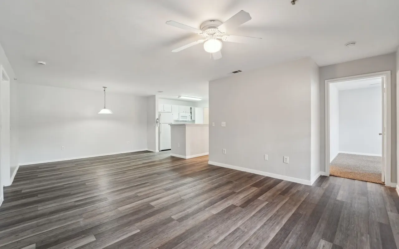 A spacious, empty living room with light-colored walls and dark wooden flooring, featuring a ceiling fan and a doorway leading to another room.