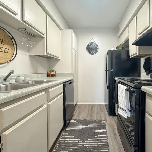 A modern kitchen featuring white cabinets, gray countertops, and black appliances. The kitchen is narrow with a rug on the floor and decorative wall art.