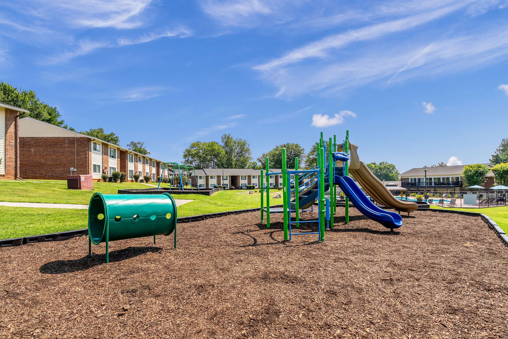 Community Playground with Apartment Views A community playground featuring a slide, climbing structure, and activity barrel, set against a backdrop of apartment buildings.