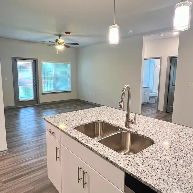 View of a modern apartment interior showcasing an open kitchen layout with a granite countertop, stainless steel sink, and two pendant lights. The living area has a ceiling fan and large windows. Doorways lead to other rooms.