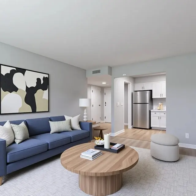 Modern living room featuring a blue sofa, round coffee table, light colors, and a view into a kitchen area.