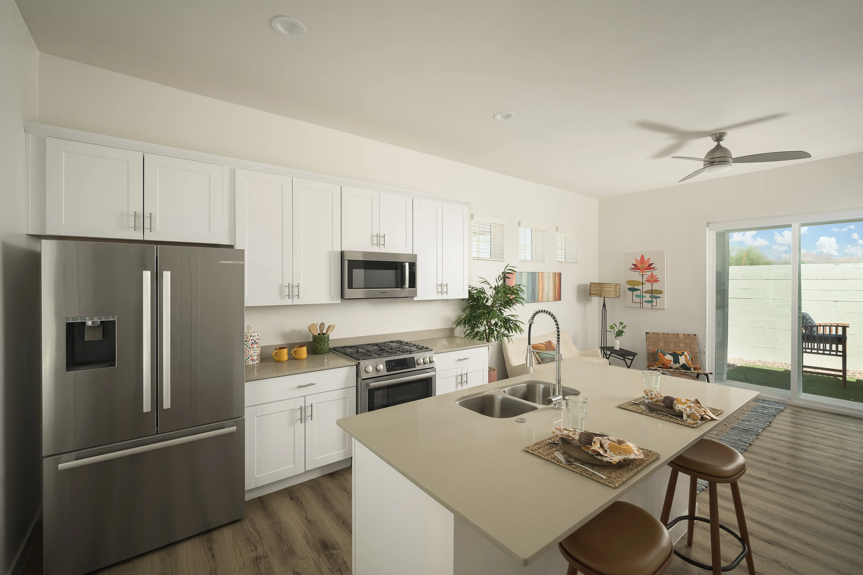 A modern kitchen featuring white cabinets, stainless steel appliances, and a large island with two bar stools. Natural light streams in from a sliding glass door leading to an outdoor patio area. Decorative items and plants are arranged on the countertop and walls.
