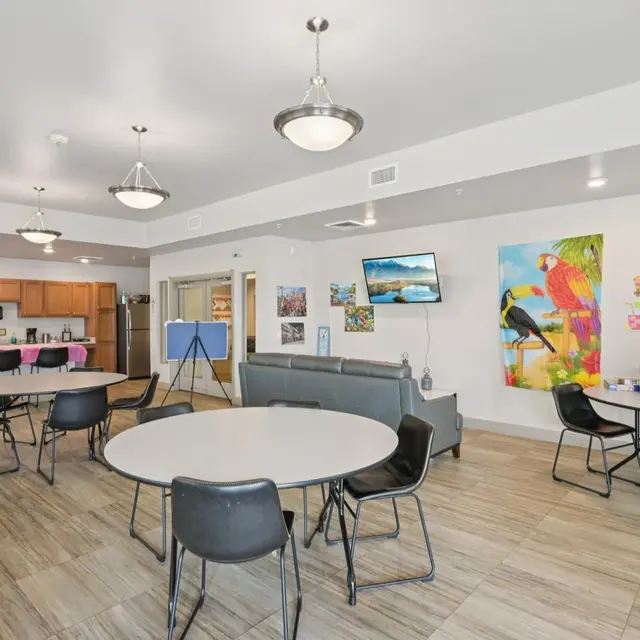 Community Room Interior A spacious community room featuring several round tables with black chairs, a kitchenette area in the background, and colorful wall decorations including paintings and posters. A TV is mounted on one wall, and there is a large gray couch in the center.